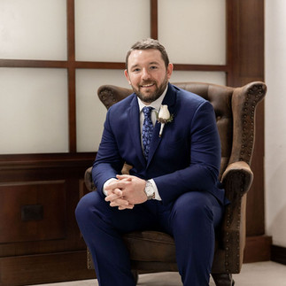Groom in navy suit sitting on a brown chair.