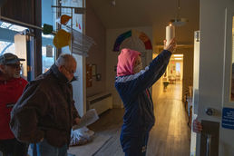 In the center of the frame, a woman wearing a hood, jacket, and medical mask raises a lit white candle in the narthex of a church building. Behind her two older men are entering the building.