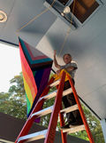 A man with a white mustache and gray-white hair stands atop a red ladder under the eave of a church building. He is holding a Progress Pride flag which he is stringing up via a pulley system