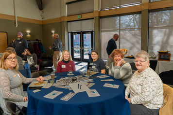 five women sitting at a round table covered in a dark blue tablecloth. Several papers are scattered around the table. A few additional people are milling around in the background.