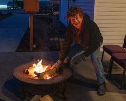 a tall woman with short hair, dressed in a coat and jeans, adds a log to a lit portable fire pit in the dim light of pre-dawn