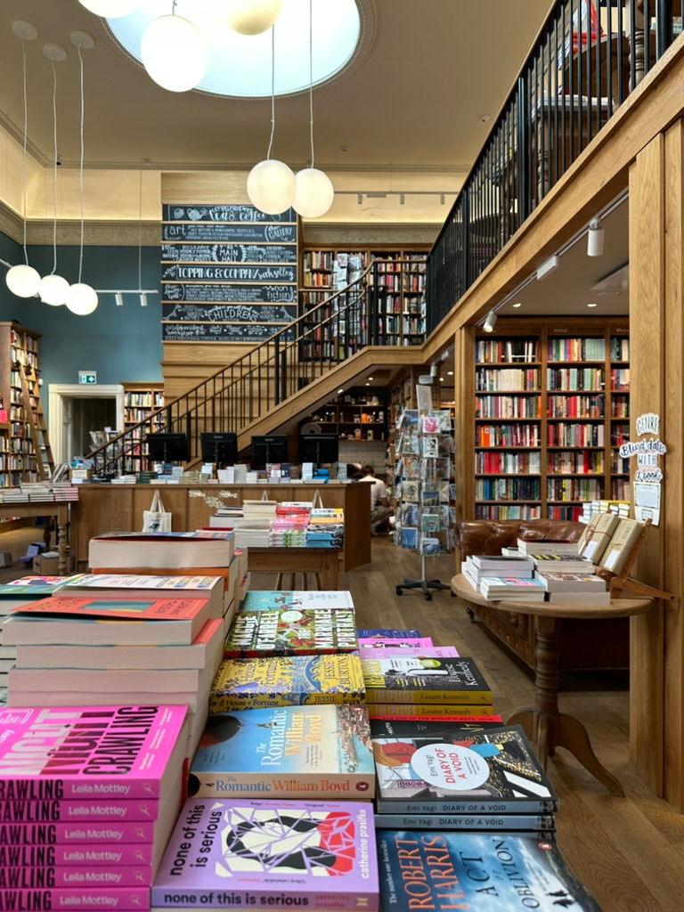 Bookstore interior with wooden shelves full of books, a staircase, colorful book displays on tables, and chalkboard signs. Bright, cozy atmosphere.