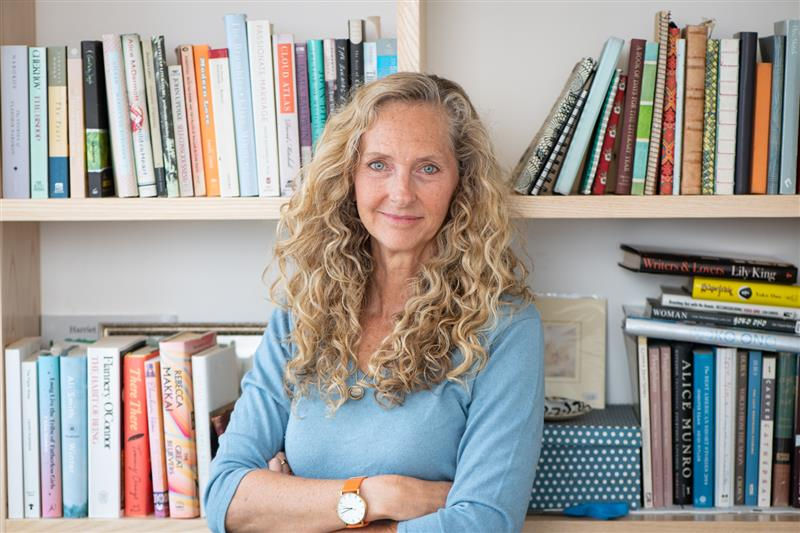 Woman with curly hair in blue sweater smiles in front of bookshelves filled with colorful books. She has an orange watch. Cozy setting.