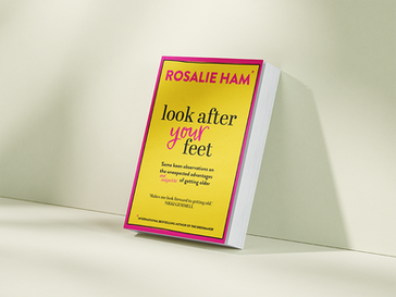 Book titled "look after your feet" by Rosalie Ham, with a yellow cover and pink accents, is leaning against a beige backdrop. Text praises book.