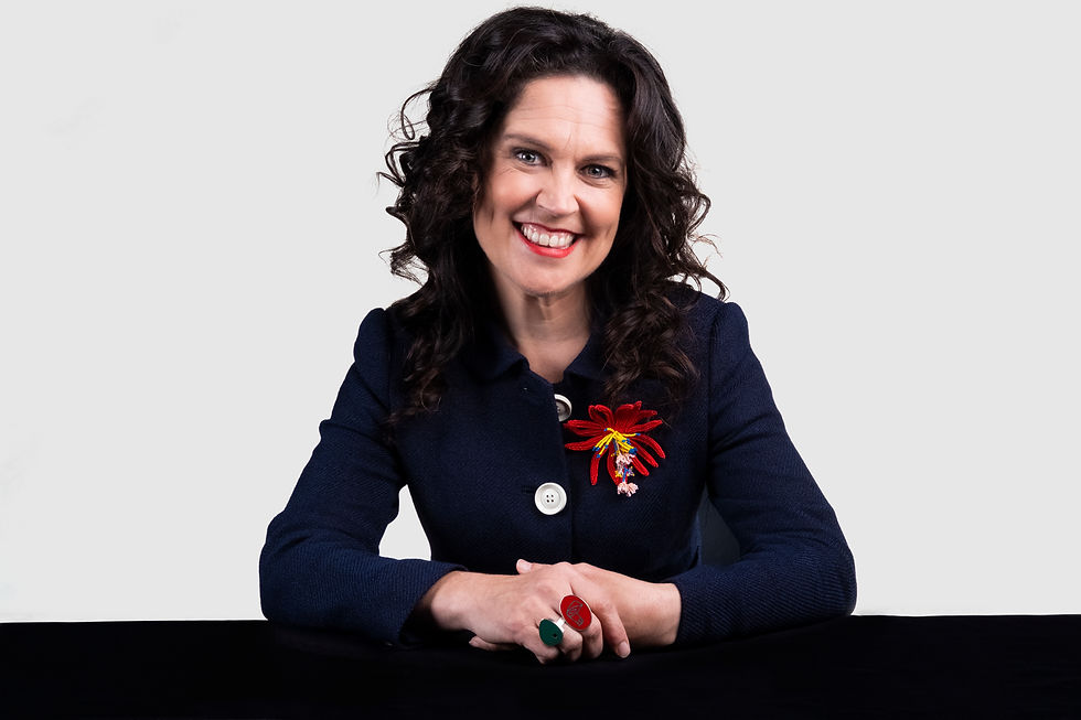 Smiling woman with curly hair in navy blazer with red flower brooch, seated at a black table against a plain white background.