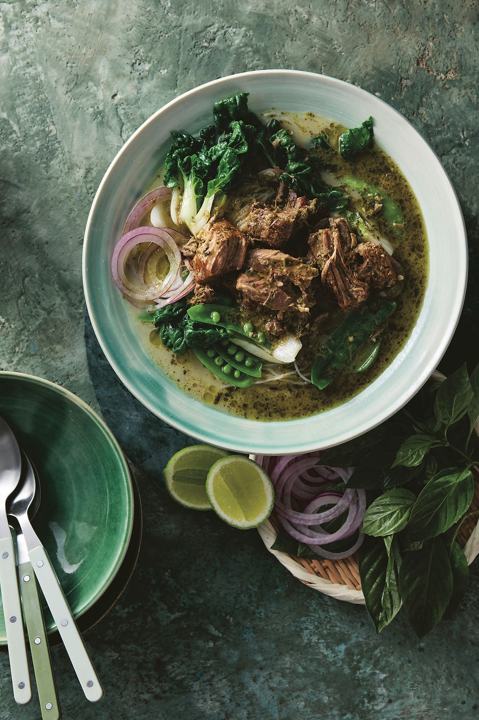Bowl of beef curry with green veggies, noodle soup, and sliced onions on a textured table. Lime halves and green leaves nearby. Cozy vibe.