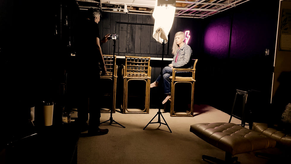Liz Short sits on a high chair under studio lights, being filmed by a person. Background has dark walls and a purple neon lightning sign. Building Connections Faster
