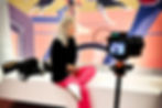 What It Takes to Scale. Liz Short in black top and pink pants sits on a bench, camera filming her. Background features colorful mural with abstract figures.