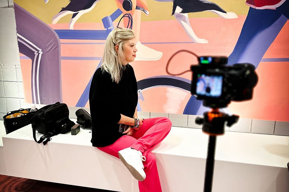 What It Takes to Scale. Liz Short in black top and pink pants sits on a bench, camera filming her. Background features colorful mural with abstract figures.