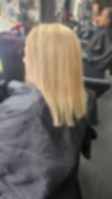 Two women in a salon, one with blonde hair facing left, another getting a haircut. Colorful wall art and hair products in the background.
