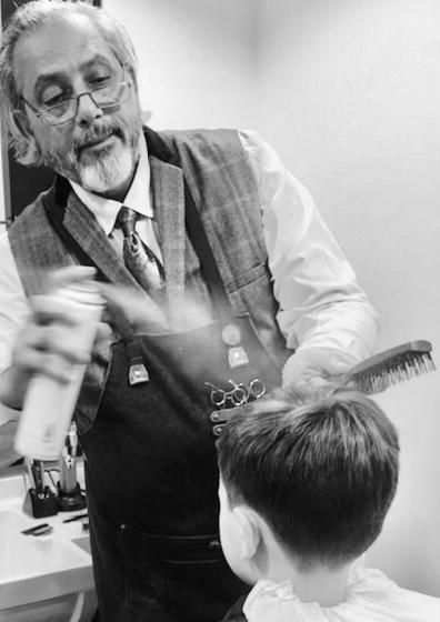 Barber sprays hair of a boy in a salon. The barber is in a vest and tie, holding a brush. The setting is calm and professional. Black and white.