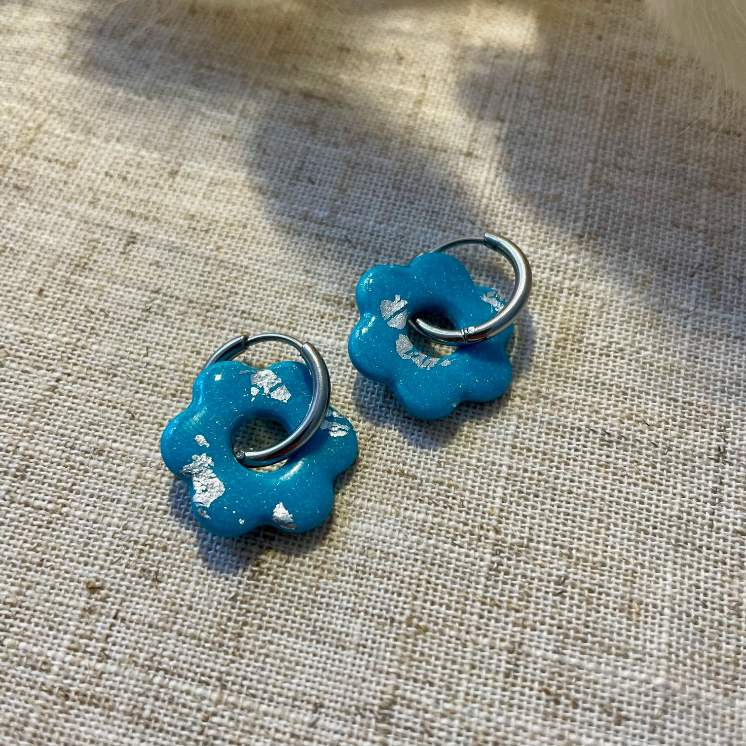 Créoles avec fleurs bleues en pâte polymère, incrustées de feuilles argentées, posées sur tissu beige