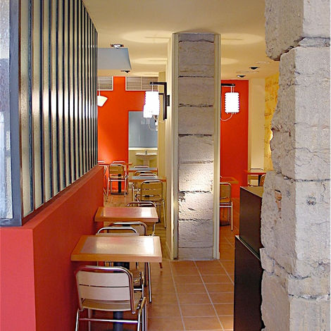 Salle de restaurant rythmée par des murs orange lumineux, un sol en terre cuite et un mobilier contemporain aux lignes épurées.
Jeu de contrastes entre pierre brute, surfaces texturées et éclairage chaleureux.