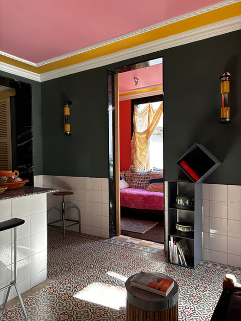 L'image représente une scène intérieure avec un design vibrant et éclectique. Le plafond est peint d'un rose frappant, bordé d'une bande jaune et d'une moulure de couronne blanche détaillée. Les murs sont principalement d'un vert foncé profond, avec une section inférieure carrelée en carrés blancs.
Le sol est recouvert d'un carrelage à motifs avec des motifs complexes dans les tons de gris, de rouge et de beige. La lumière du soleil s'écoule d'une source inaute, projetant des ombres géométriques sur le sol, en particulier au premier plan.
Sur le côté gauche de l'image, une partie d'un comptoir de cuisine est visible. Il a une façade carrelée blanche et un comptoir moucheté foncé. Un bol rouge avec quelques éléments décoratifs se trouve sur le comptoir. À gauche du comptoir, un panneau effet marbre noir est monté. Un simple tabouret métallique avec un siège en bois est positionné près du comptoir.
Au centre de l'image, une porte ouverte mène à une autre pièce. Cette porte a un cadre effet marbre noir avec un bord intérieur en bois. La pièce au-delà est baignée d'une chaude lumière orange rougeâtre. Un lit de repos recouvert d'un tissu texturé rose vif est visible, orné de nombreux oreillers à motifs en bleu et blanc. Un rideau jaune doré est suspendu à la fenêtre de cette pièce. Le plafond de cette pièce intérieure est également rose, reflétant la couleur du plafond de la pièce principale.
À droite de la porte, une étagère moderne et grise se trouve contre le mur vert foncé. Il a plusieurs compartiments ouverts contenant des bols décoratifs et des livres. Le haut de l'étagère est incliné et a un carré noir avec un objet rouge à l'intérieur, peut-être un livre ou un objet décoratif. Deux appliques murales cylindriques avec des détails jaunes et argentés sont montées sur le mur vert foncé, une de chaque côté de la porte.
Au premier plan, un petit tabouret rond avec une base en bambou soutient un livre avec une couverture colorée. L'impression générale est celle d'un design intérieur audacieux et non conventionnel, caractérisé par de forts contrastes de couleurs et un mélange de styles.