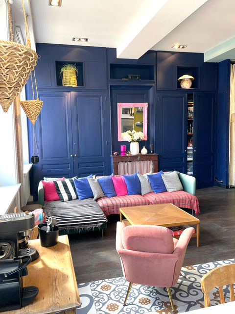 Salon avec rangements bleus et canapé multicolore
• Mobilier :
• Canapé bas en L très coloré (bleu, rose, rayures, carreaux) avec coussins contrastés.
• Fauteuil rose poudré en velours.
• Table basse en bois clair, design sobre.
• Mur : Grande bibliothèque/rangement sur mesure bleu nuit, miroir rose vif au centre.
• Style : Mélange de chic pop, vintage et design coloré.