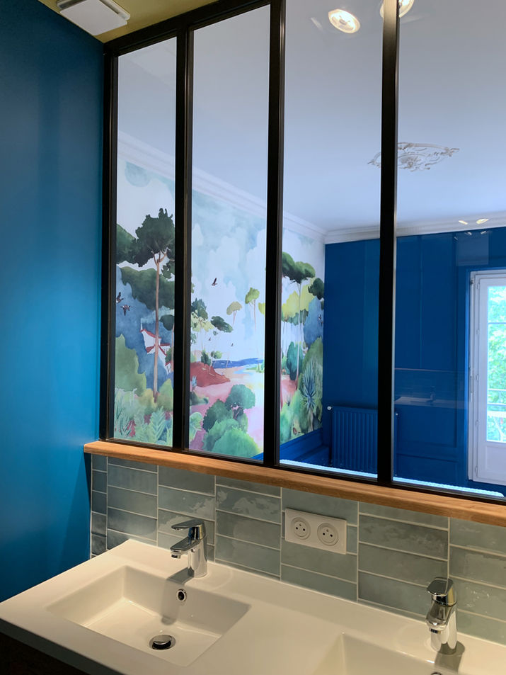Premier plan :
	•	Double vasque blanche moderne avec robinetterie chromée.
	•	Crédence en carreaux vert d’eau posés à l’horizontale, finition brillante.
	•	Élément marquant :
	•	Verrière intérieure noire de type atelier, séparant la salle de bain d’une autre pièce tout en laissant passer la lumière.
	•	En arrière-plan :
	•	Magnifique fresque murale représentant un paysage naturel et coloré, type aquarelle, avec arbres, végétation et vue sur la mer.
	•	Mur bleu profond et grande fenêtre visible à droite.
	•	Ambiance : Fraîche, artistique et lumineuse. Très contemporaine, mais avec un vrai caractère poétique.