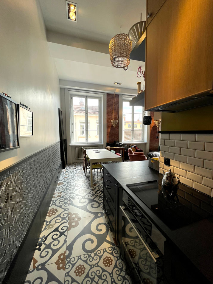  Vue de la cuisine vers le salon
	•	Sol : Carreaux de ciment graphiques, mélange de courbes, rosaces et couleurs douces (gris, bleu, ocre).
	•	Cuisine : Mobilier bas noir mat, plan de travail noir, crédence en carreaux métro blancs.
	•	Mur : Partie haute blanche, partie basse à motifs géométriques gris et noir.
	•	Éclairage : Suspensions en osier, ambiance bohème.
	•	Au fond : Espace salon avec lumière naturelle, murs en briques rouges apparentes, grande fenêtre, mobilier vintage.