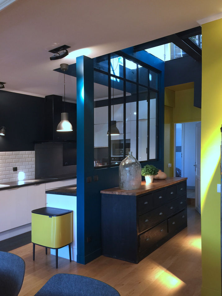 
Cette photographie d'intérieur présente un espace de vie moderne et bien aménagé, caractérisé par des couleurs vives et une conception architecturale distinctive.

La composition de la scène révèle une séparation entre deux espaces : une cuisine et un autre espace non clairement défini, probablement un salon ou une salle à manger. Une cloison en acier noir et verre, peinte en bleu foncé, sert de séparation, permettant à la lumière naturelle de circuler tout en délimitant les zones. Un meuble bas noir avec un dessus en bois clair est placé devant la cloison, accueillant des objets décoratifs. La perspective est prise de manière à mettre en valeur les lignes épurées et la palette de couleurs audacieuse.

Les éléments clés incluent le meuble bas en bois et noir, orné d'une grande jarre en verre et d'une plante verte, et un seau à déchets jaune vif. La cloison en verre et métal noir sépare visuellement les espaces, tandis qu'une cuisine aux murs blancs et noirs se distingue à l'arrière-plan. Les luminaires suspendus ajoutent une touche industrielle moderne. Le choix des couleurs vives crée un contraste intéressant avec les tons neutres des murs et du mobilier.

La photographie capture un style contemporain, avec une utilisation prononcée de la lumière naturelle et des contrastes de couleurs. L'esthétique met en avant les matériaux nobles, comme le bois et le verre. La profondeur est créée par la perspective, et l'attention aux détails architecturaux révèle une approche soignée de l'aménagement intérieur. Le choix des couleurs vives, le jaune et le bleu, dynamisent l'espace et reflètent un design moderne.

L'environnement est baigné de lumière naturelle, probablement provenant d'une grande fenêtre ou d'une source de lumière extérieure non visible. L'éclairage met en valeur les différents éléments décoratifs et les matériaux. L'atmosphère est à la fois accueillante et élégante, suggérant un espace de vie soigné et fonctionnel. Le sol en bois clair renforce la sensation de chaleur et de convivialité, tandis que les couleurs vives ajoutent une touche de dynamisme et de modernité.

Rénovation d'un duplex dans un esprit loft industriel.
Notre réflexion a principalement consisté à donner une "âme " à ces beaux volumes existants en insufflant un esprit loft industriel.
Nous avons invité la lumière naturelle et artificielle à se frayer un chemin par la création de verrières, d'un plancher de verre et le positionnement de miroirs. 
Les matériaux bruts viennent en contraste avec des compositions subtiles de tapisseries ou le raffinement de moulures en staff.
Quant à la couleur, elle joue son rôle d'accentuation des volumes dans une palette riche en tonalité et contraste.