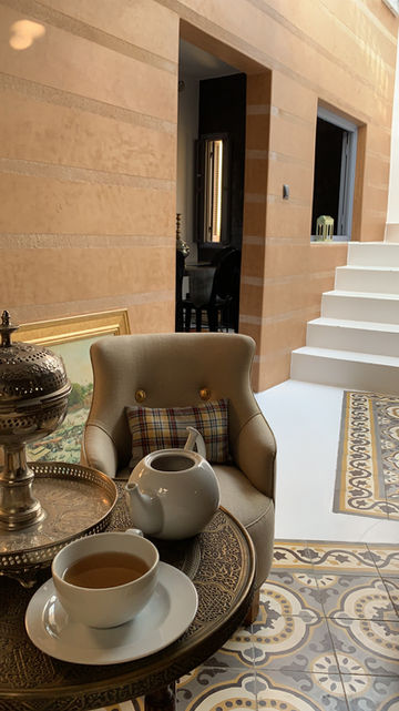 L'image capture une scène intérieure sereine et élégante, probablement un salon ou un patio dans une maison moderne ou un établissement de luxe. La composition est équilibrée et accueillante, en mettant l'accent sur le confort et l'esthétique raffinée.
Au premier plan, un fauteuil rembourré beige en peluche avec un dossier capitonné et deux boutons dorés décoratifs est bien en vue. Repose contre le dossier de la chaise se trouve un coussin à carreaux avec un motif à carreaux classique dans les tons de beige, marron et rouge sourd. Sur un plateau rond en laiton orné placé devant la chaise se trouve une théière en céramique blanche avec un bec et une poignée incurvés, remplie d'un liquide sombre, probablement du thé. À côté, le bord d'une tasse de thé blanche assortie est visible.
À gauche de la chaise, un grand brûleur d'encens argenté au design complexe avec des détails perforés repose sur le même plateau en laiton. Derrière la chaise, partiellement visible, se trouve une peinture encadrée avec une bordure dorée ; l'œuvre d'art représente une scène vivante, peut-être historique ou rurale, avec de la verdure et des figures, ajoutant une touche de charme classique.
L'arrière-plan révèle un espace intérieur aux tons chauds. Les murs sont finis dans une couleur lisse, terre cuite ou ocre clair avec des bandes horizontales. À droite, un ensemble d'escaliers blancs propres monte vers une porte ou un niveau supérieur. À travers la porte, on peut voir un aperçu d'une salle à manger avec des meubles en bois sombre. Sur le mur près des escaliers, une petite lanterne verte décorative se trouve sur un rebord.
Le sol est carrelé avec une base blanche et présente une bordure décorative au premier plan, présentant un motif géométrique et floral complexe dans des tons terreux de beige, brun et or. L'éclairage général est doux et naturel, suggérant la journée, et la palette de couleurs est chaude et harmonieuse, dominée par des accents beige, crème, terre cuite et métalliques. La scène transmet un sentiment de calme, de luxe et de design réfléchi.