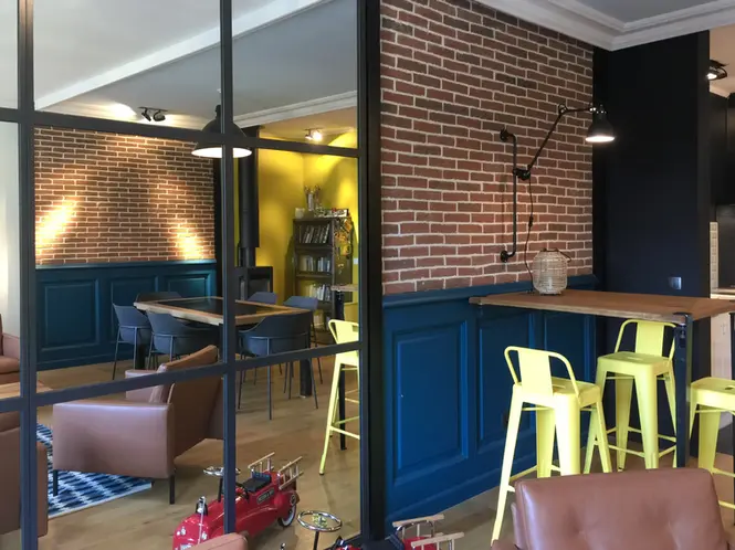 Aménagement d’une cuisine ouverte mêlant verrière atelier, mur en briques apparentes, bar en bois sur mesure, assises colorées et palette contrastée pour créer un intérieur au caractère industriel chaleureux.