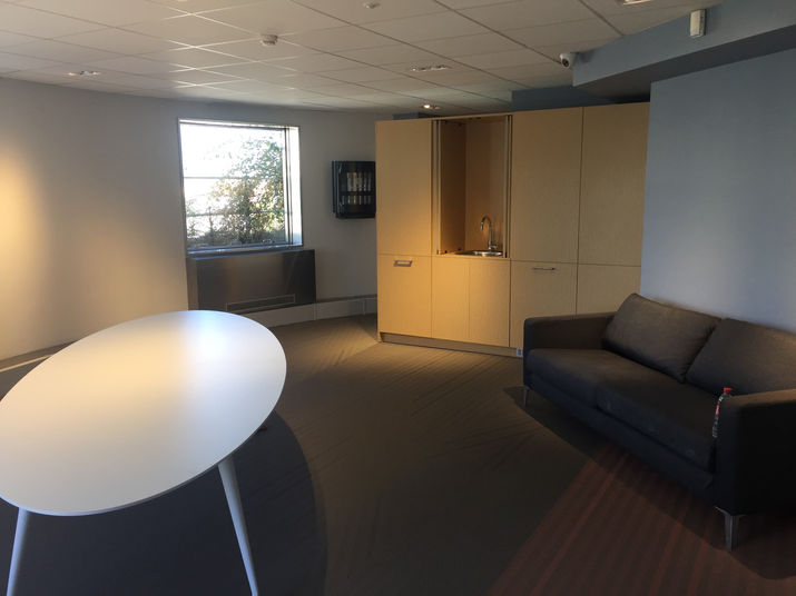 L'image représente un espace intérieur moderne et minimaliste, probablement une salle de pause ou un salon dans un bureau ou un bâtiment institutionnel. La pièce est bien éclairée avec une lumière naturelle qui entre d'une grande fenêtre rectangulaire sur le mur de gauche, qui révèle un feuillage vert à l'extérieur. La fenêtre est située au-dessus d'une plinthe métallique foncé ou d'une unité de chauffage.

Au premier plan, une grande table blanche de forme ovale avec de fins pieds blancs occupe le côté gauche du cadre, sa surface reflétant la lumière ambiante. À droite, un canapé trois places gris foncé ou noir avec un profil bas et des pieds métalliques fins repose contre le mur. Une seule bouteille d'eau en plastique avec un bouchon rouge est placée sur le côté droit du canapé.

Derrière le canapé, une grande armoire de couleur bois clair s'étend sur le mur. L'une des portes de l'armoire est ouverte, révélant un petit évier avec un robinet chromé à l'intérieur. L'armoire a un design épuré et moderne avec des poignées horizontales simples. Au-dessus de l'armoire, monté sur le mur, se trouve un dispositif rectangulaire noir qui semble être un distributeur automatique ou une unité de rangement murale.

Le sol est recouvert d'un tapis sombre et texturé avec un motif subtil, peut-être un tissage à chevrons ou diagonal. Le plafond est composé de carreaux acoustiques blancs disposés dans un motif de grille, avec des luminaires encastrés et au moins une caméra de sécurité visible montée près du coin supérieur droit. Les murs sont peints dans une couleur gris clair neutre ou blanc cassé, contribuant à l'esthétique calme et fonctionnelle de la pièce. La composition globale est équilibrée, la table et le canapé créant un sentiment de symétrie, tandis que l'armoire ouverte et la fenêtre offrent un intérêt visuel et de la profondeur.