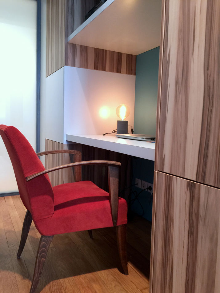 Bureau intégré entre des colonnes de rangements, mélangeant bois veiné clair, blanc mat, et fond bleu.
	•	Chaise vintage tapissée de velours rouge, apportant chaleur et contraste à l’espace.
	•	Petite lampe à ampoule Edison, ajoutant une touche rétro et une lumière douce.