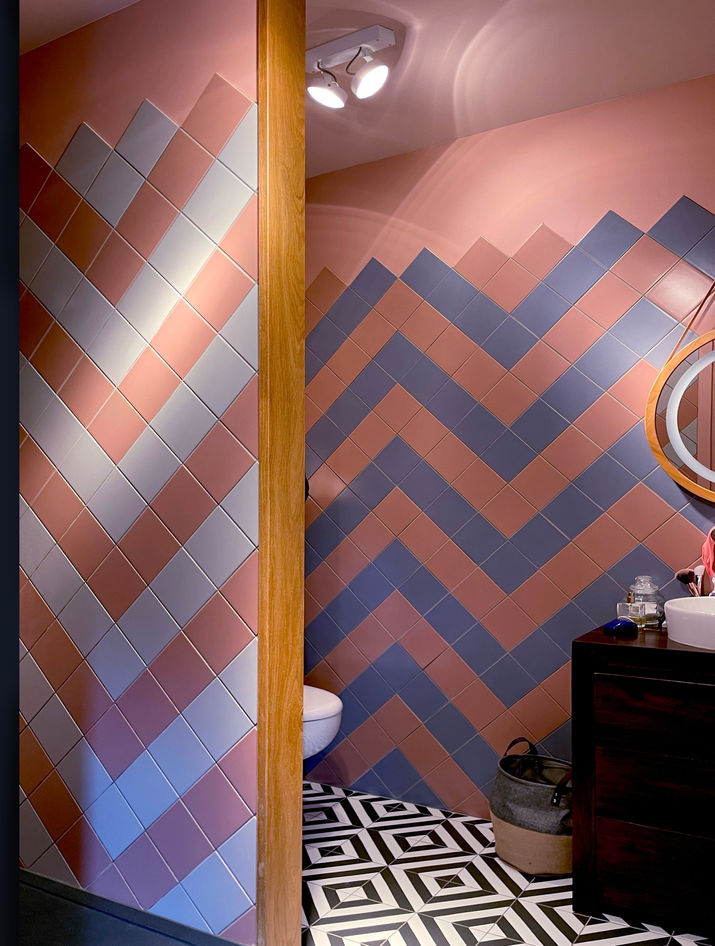 Salle de bain originale en rose et bleu
	•	Style : Audacieux et graphique.
	•	Éléments clés :
	•	Murs en carrelage rose, bleu et blanc posés en motifs en chevron et en diagonale.
	•	Sol en carrelage à motif 3D en noir et blanc, très fort visuellement.
	•	Meuble vasque en bois foncé avec vasque blanche.
	•	Décoration éclectique et très personnelle.