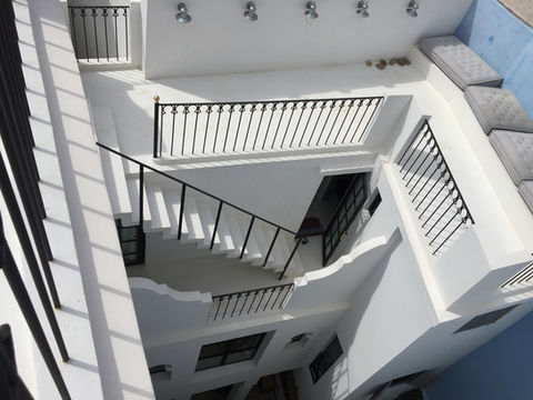L'image montre une cour intérieure à plusieurs niveaux et un escalier blanc proéminent. L'architecture est propre et moderne, avec des murs et des plafonds blancs éclairés par la lumière naturelle. L'escalier lui-même est également blanc, avec une balustrade en métal noir qui présente un motif décoratif de barres verticales et d'éléments incurvés. La balustrade est surmontée d'une fine main courante noire.
La composition est d'un point de vue élevé, regardant vers le bas dans la cour. Cette perspective met l'accent sur la verticalité de la structure et la profondeur de l'espace. Des ombres sont projetées par la balustrade et l'escalier sur les surfaces blanches, créant un fort contraste et ajoutant un sentiment de dimension.
Au niveau supérieur, au-dessus de l'escalier principal, il y a un balcon avec une balustrade noire similaire. Plusieurs luminaires ronds et argentés sont fixés au plafond dans cette zone. À droite, sur le mur du haut, un petit groupe de pierres est visible.
L'escalier descend dans une série de virages. Les marches sont larges et semblent être en béton ou en un matériau similaire. La balustrade noire suit le chemin des escaliers, offrant un sentiment de guidage et de sécurité.
Sur un niveau inférieur, à droite de l'escalier, il y a une porte ouverte menant à un espace intérieur plus sombre. Une fenêtre avec des cadres sombres et des vitres est visible à l'intérieur de cette ouverture. Une caméra de sécurité est montée sur le mur près de la porte.
Plus bas, une autre section de garde-corps noir est visible, indiquant encore un autre niveau du bâtiment. Sous cette balustrade, une fenêtre rectangulaire avec un cadre sombre est serrée dans un mur blanc. D'autres fenêtres et détails architecturaux sont suggérés dans les parties inférieures de l'image, suggérant un bâtiment à plusieurs étages avec une zone centrale ouverte.
La palette de couleurs est dominée par le blanc, avec des accents de noir sur les balustrades et les cadres de fenêtres. L'éclairage est lumineux et naturel, créant une atmosphère propre et aérée. L'impression générale est celle de l'élégance minimaliste et du design architectural.