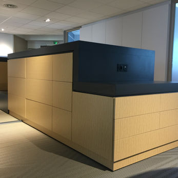 L'image montre une réception de bureau moderne. Le bureau est en forme de L et semble être en bois de couleur claire pour le corps principal, avec un comptoir et une partie supérieure gris foncé ou noir. Les panneaux en bois à l'avant du bureau sont disposés dans une grille de tiroirs, avec trois tiroirs visibles sur la partie avant et plus de tiroirs à gauche. Le grain du bois est subtil et court verticalement sur les façades des tiroirs visibles. La partie supérieure sombre a un design épuré et angulaire et contient deux prises électriques. L'impression générale est celle du design et de la fonctionnalité contemporains.

La scène est l'intérieur d'un bureau, probablement un hall ou une zone de réception. Le revêtement de sol est recouvert d'un motif géométrique de rayures gris clair et blanches. Le plafond est un plafond tombant avec des carreaux carrés et des appareils d'éclairage encastrés. En arrière-plan, à travers une porte ouverte à droite, un espace de bureau est visible avec un bureau et une chaise de bureau. L'éclairage dans l'image est un mélange de plafonniers ambiants et peut-être de lumière naturelle provenant des fenêtres, créant des ombres douces.

Les couleurs sont principalement neutres, les tons chauds du bois contrastant avec le gris froid et le noir du bureau et du revêtement de sol. L'éclairage projette une lueur chaude sur les surfaces en bois. La composition place le bureau bien en évidence au premier plan, remplissant une partie importante du cadre, avec les éléments d'arrière-plan légèrement flous, attirant l'attention sur le sujet principal. La perspective est d'un angle légèrement bas, en regardant le bureau. Les lignes du bureau sont fortes et rectilignes, soulignant son esthétique moderne.