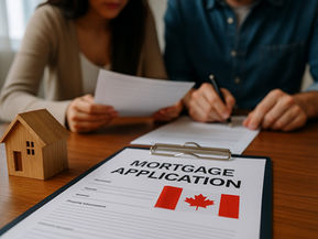 A close-up view of a Canadian mortgage application form on a clipboard with a red maple leaf flag, next to a small wooden house model, while a couple in the background reviews financial documents at a wooden table in a home setting.