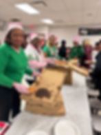 Alpha Gamma Omicron Omega Chapter members dressed in pink and green AKA sweatshirts with pink Santa Claus hats, handing out pizza to young teens at a community event.