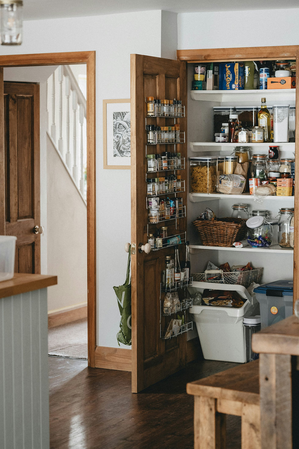 Open pantry with jars, spices, pasta, and snacks. Wooden shelves, white walls, and a staircase visible. Cozy kitchen setting.