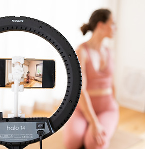 een ringlamp rond een smartphone terwijl iemand in sportkledij op de achtergrond zit, alsof er fitnesscontent wordt opgenomen