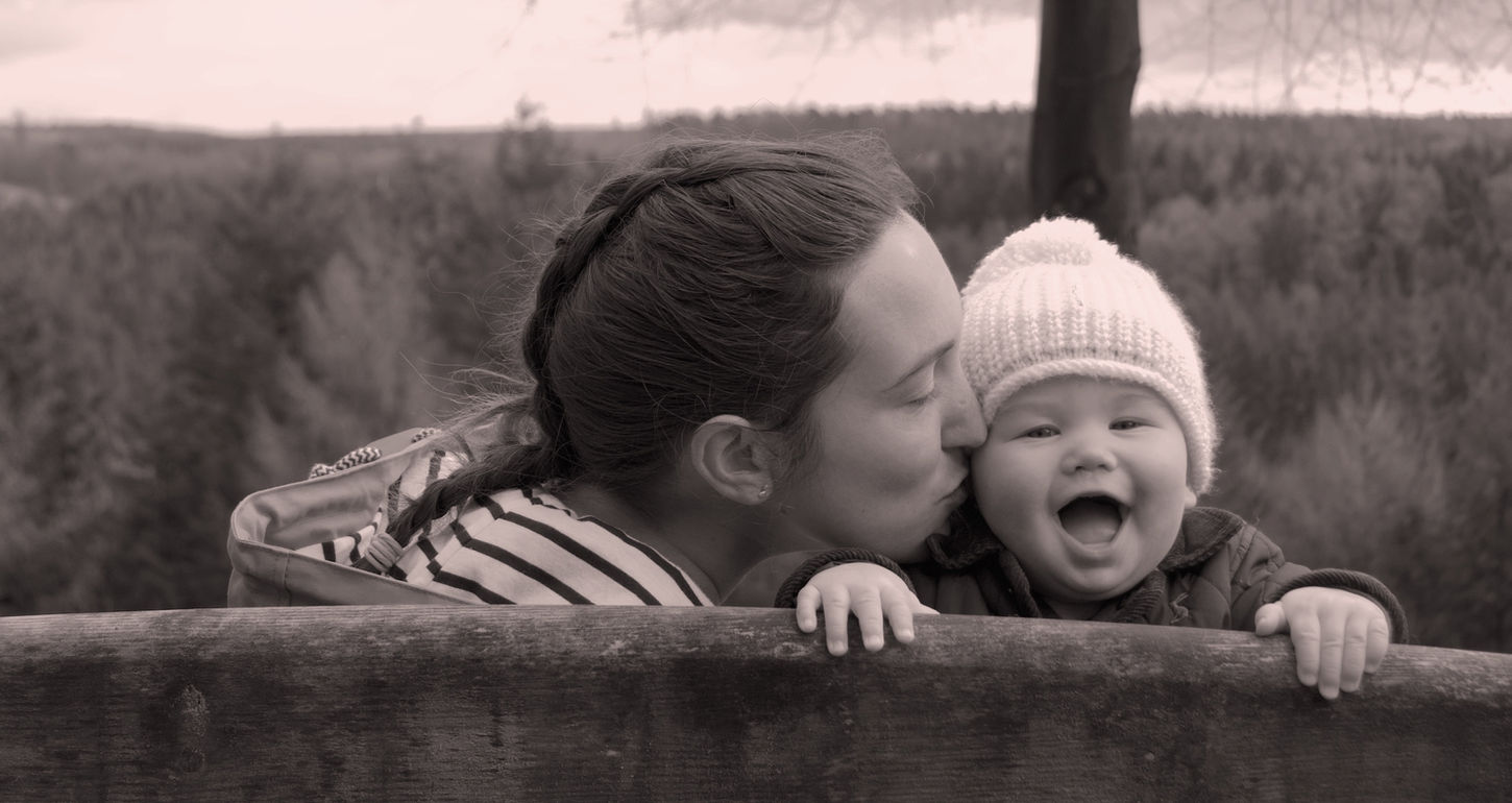 A black and white photo of a mother with a french plait in her hair kissing her baby on the cheek. The baby is laughing towards the camera, over the back of a bench.