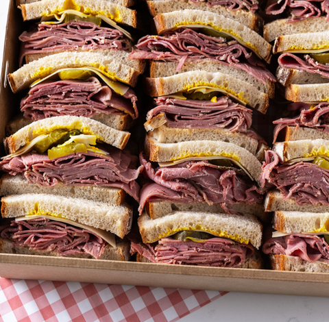 Platter of sliced pastrami on rye sandwiches with mustard and pickles, neatly arranged on a serving tray.