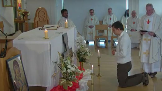 A person kneels in prayer before an altar with candles and flowers. Four seated clergy in white robes and one standing read from a book.