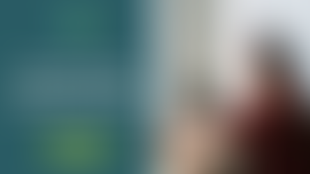 Woman in red patterned shirt wearing headphones, working on a laptop by a window.