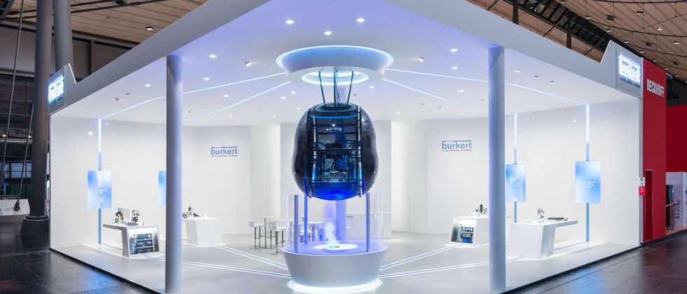 Bürkert Messestand : Foto der offenen Standfront mit schwebender transparenter Kapsel, blauen Lichtakzenten und weißen Produktstationen.