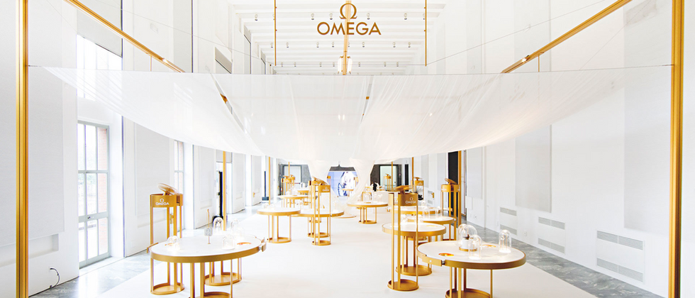 Omega - Her Time: Foto der hellen Uhrenausstellung mit goldfarbenem Rahmen, runden Präsentationstischen und schwebenden weißen Stoffbahnen.