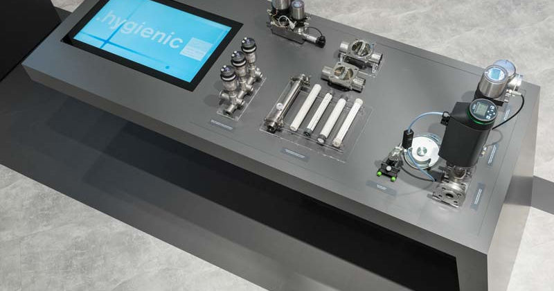 Bürkert Messestand : Nahansicht eines schwarzen Ausstellungstischs mit Bildschirm und metallischen technischen Komponenten.