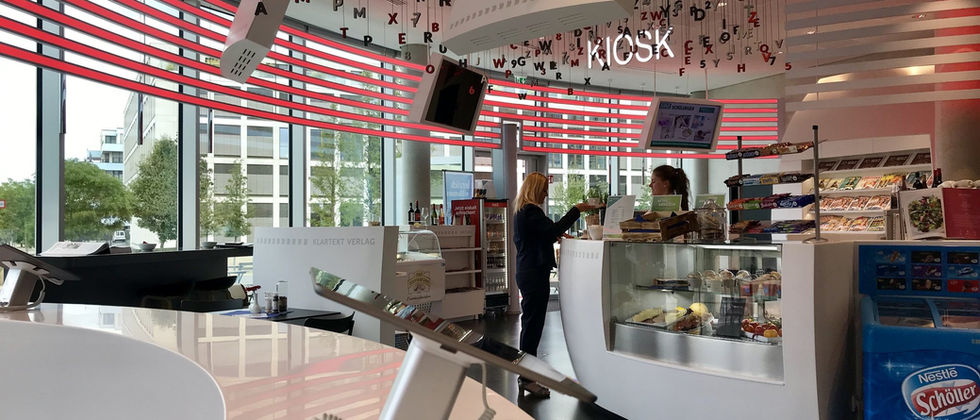 Funke Medien Gruppe- Kiosk: Foto der weißen gebogenen Theken mit roten horizontalen Lichtakzenten und einer Buchstabeninstallation an der Decke.