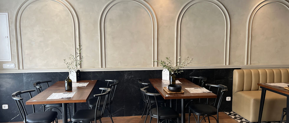 Mustache Restaurant : Drei Holztische mit schwarzen Stühlen vor einer hellen Wand mit großen Bogenprofilen.