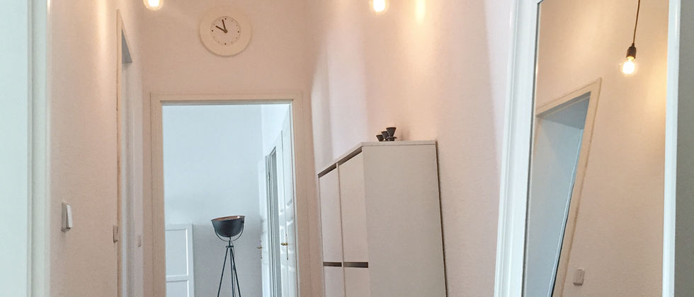 Wohnung Berlin Mitte: Foto des weißen Flurs mit Holzdielen, großem Spiegel, schmalen Schränken und frei abgehängten Glühlampen.