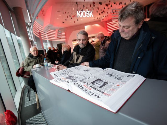 Kiosk- und Besucherbereichsdesign – FUNKE-Kiosk in Essen: Foto von Besuchern beim Zeitungslesen an einer hellen Theke unter dem Kiosk-Schriftzug und hängenden Buchstaben. | EN: FUNKE Kiosk, Essen: visitors reading newspapers at a pale counter beneath the Kiosk sign and suspended letters.