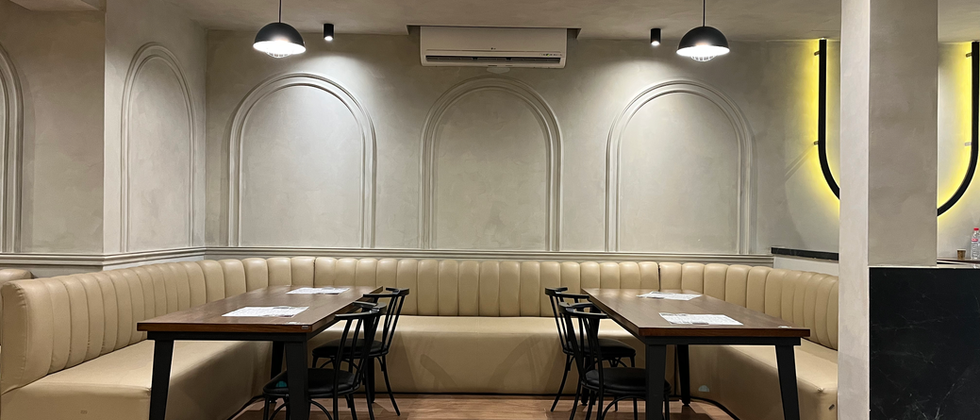 Mustache Restaurant : Beige Polsterbank mit Holztischen und schwarzen Pendelleuchten vor heller Wand mit Bogenprofilen.