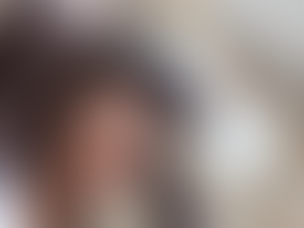 Woman cheerfully holding books on her head, smiling broadly. Background shows blurred building exterior with light-colored windows.