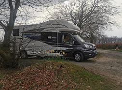 Black motorhome for rent, parked in a natural setting with bare trees.