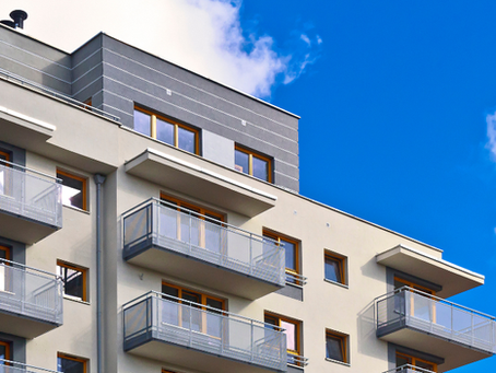 Prédio residencial moderno com varandas de vidro sob céu azul com nuvens brancas.