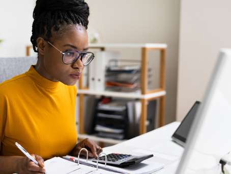 Mulher de óculos e blusa amarela trabalha no escritório, anotando e usando calculadora diante do computador.
