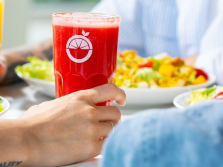 Mão segura copo vermelho de suco com logo de laranja, à mesa com saladas coloridas ao fundo.
